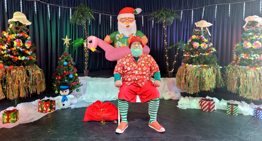 Santa Kenny sitting on lawn chair surrounded by Christmas trees decorated with leis